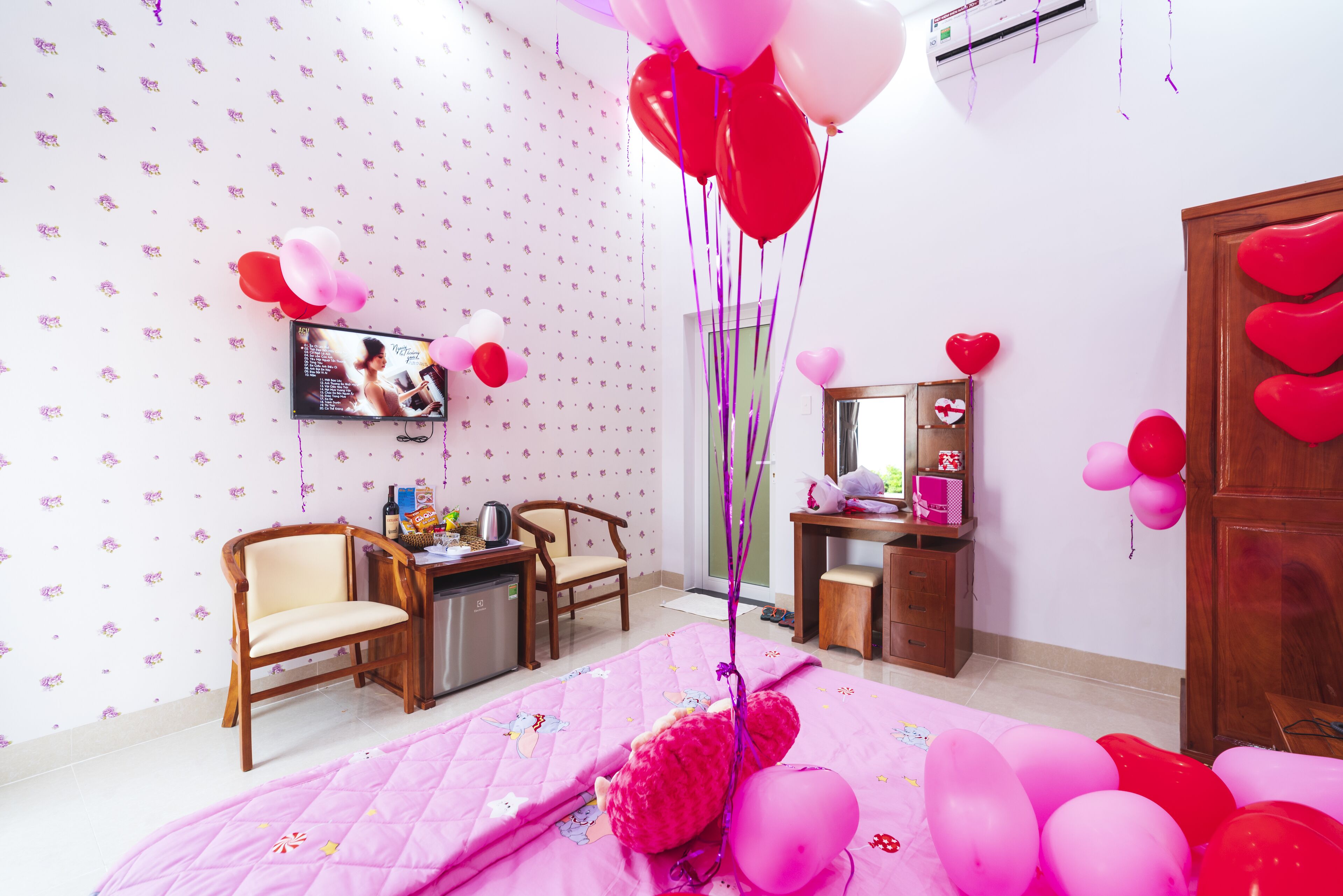 Quarto com uma colcha rosa, cadeiras de madeira, uma escrivaninha com computador e uma televisão na parede.