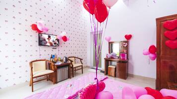 Quarto com uma colcha rosa, cadeiras de madeira, uma escrivaninha com computador e uma televisão na parede.
