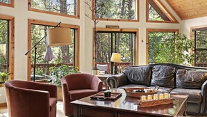A spacious living room with a leather sofa, two armchairs, and a coffee table with a tray of glasses and a book. Large windows offer a view of the surrounding trees. - Chalet Forest Views, 2 kings, fireplace, decks (Pine)