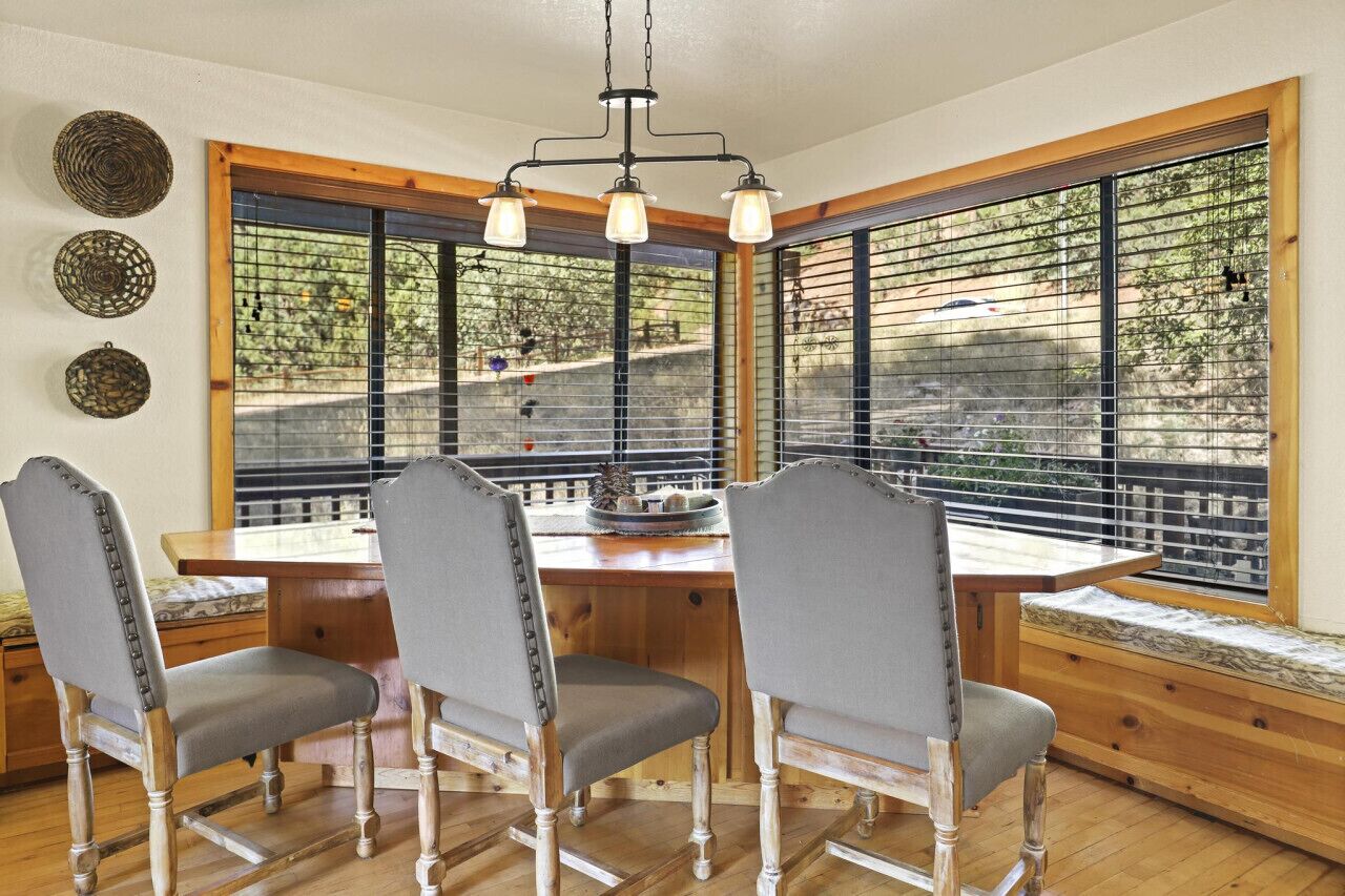 A dining area with a wooden table and four chairs, large windows with blinds, and a hanging light fixture.