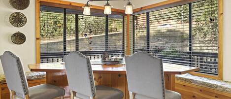 A dining area with a wooden table and four chairs, large windows with blinds, and a hanging light fixture.