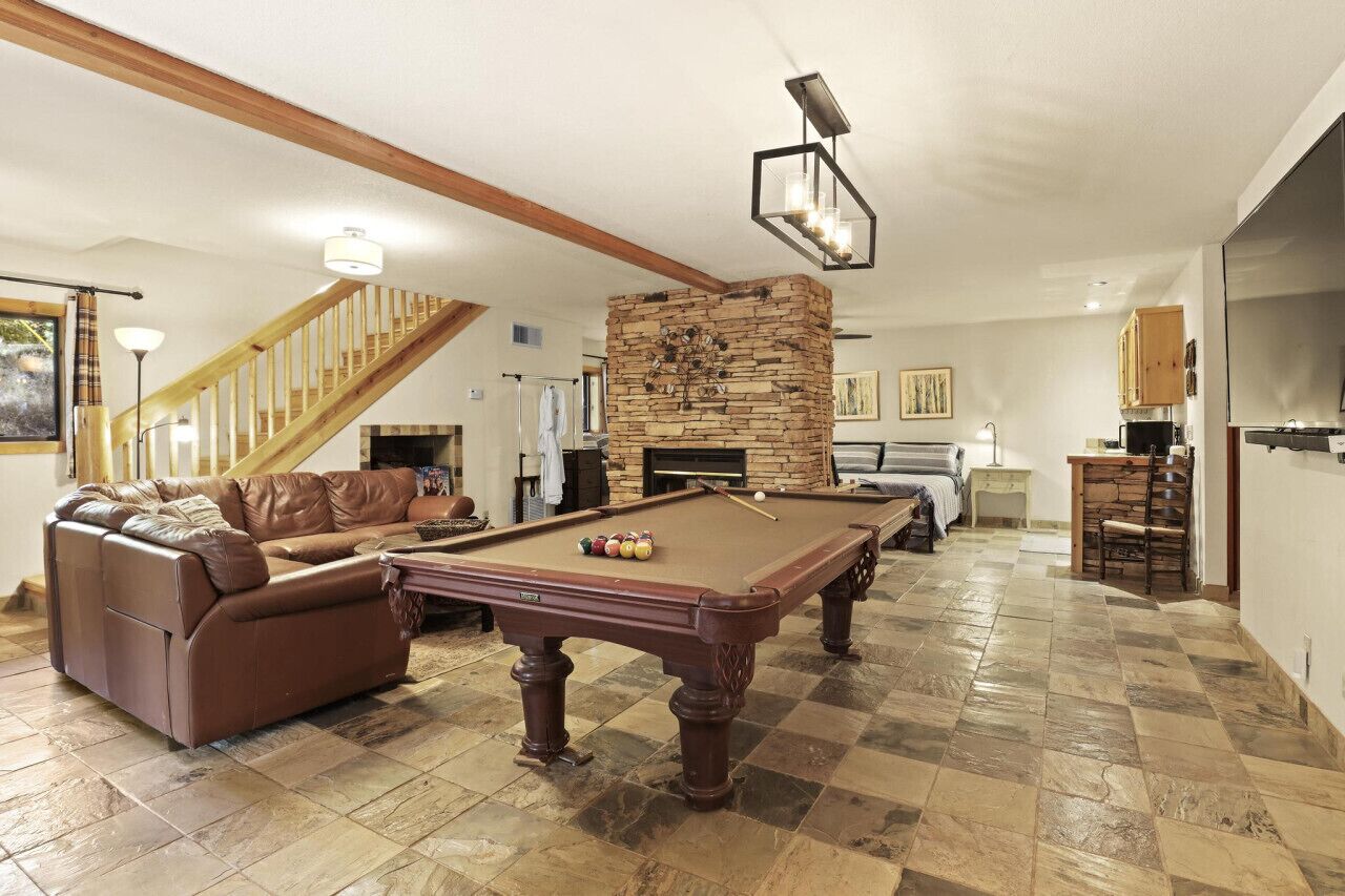 A spacious living room with a pool table, a brown leather sofa, a stone fireplace, and a staircase.
