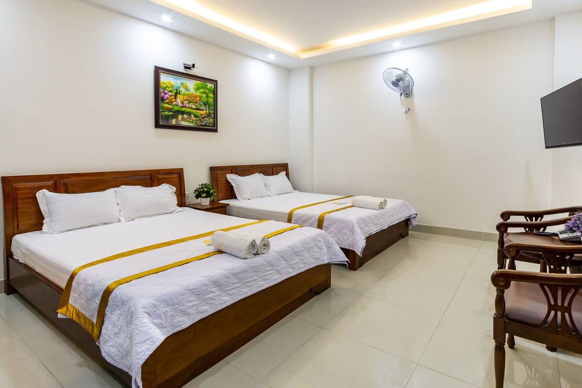 A hotel room with two beds, a TV, a fan, and a painting on the wall.