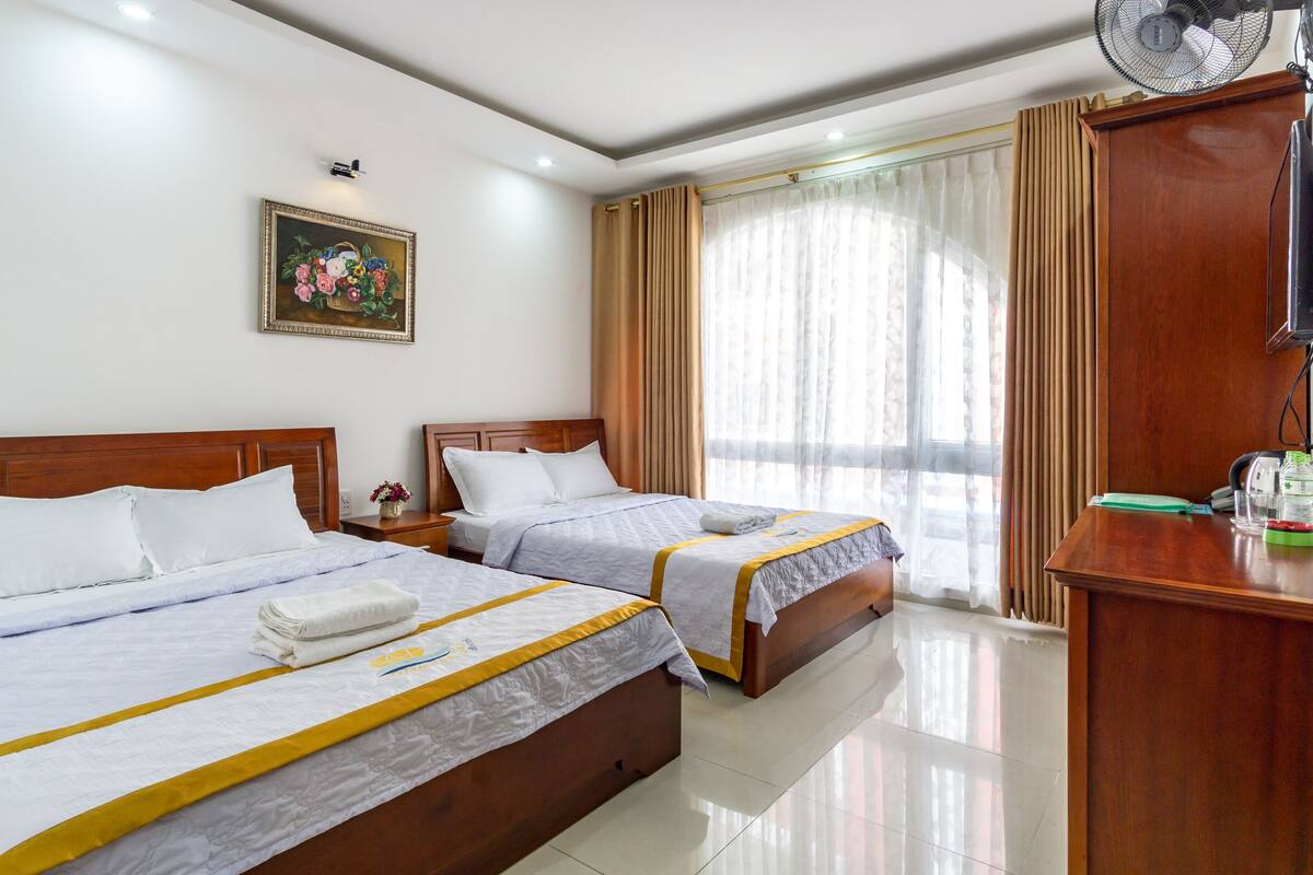 A hotel room with two beds, a wooden headboard, a desk with a computer, a fan, and a window with curtains.