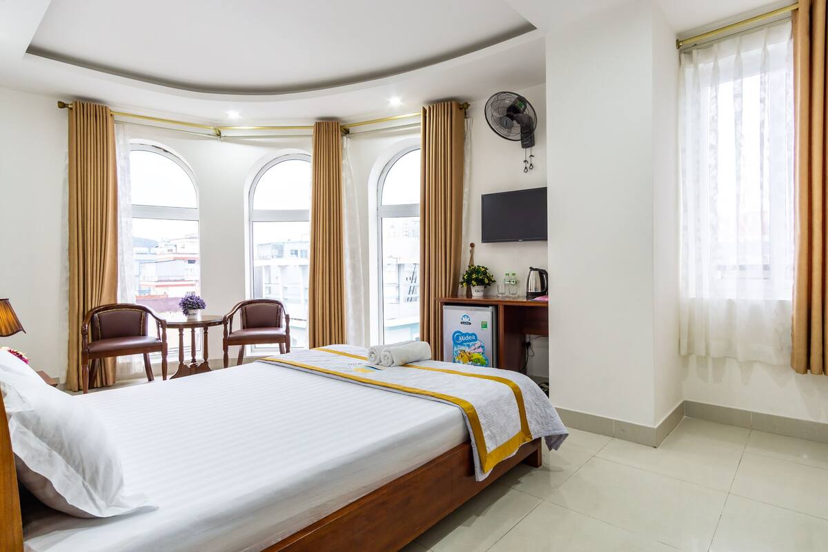 A hotel room with a large bed, two chairs, a small table, a TV, a fan, and a minibar.