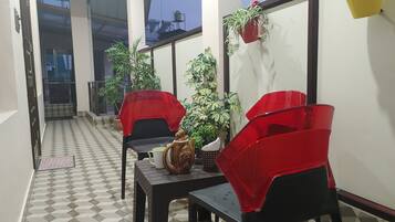 A covered outdoor seating area with two tables and four red chairs, surrounded by potted plants and a tiled floor.