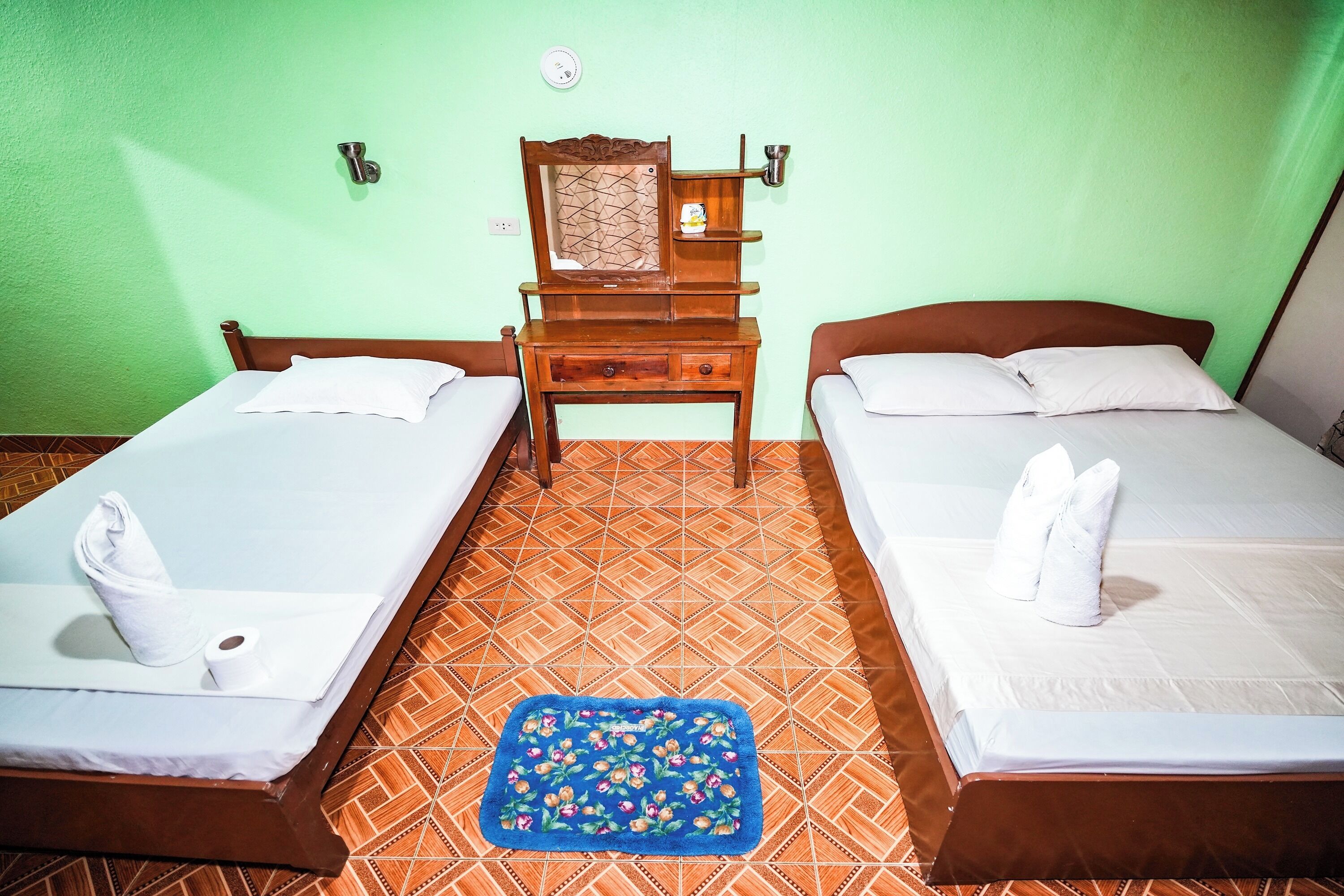 A room with two beds, a wooden nightstand with a mirror, and a blue floral rug.