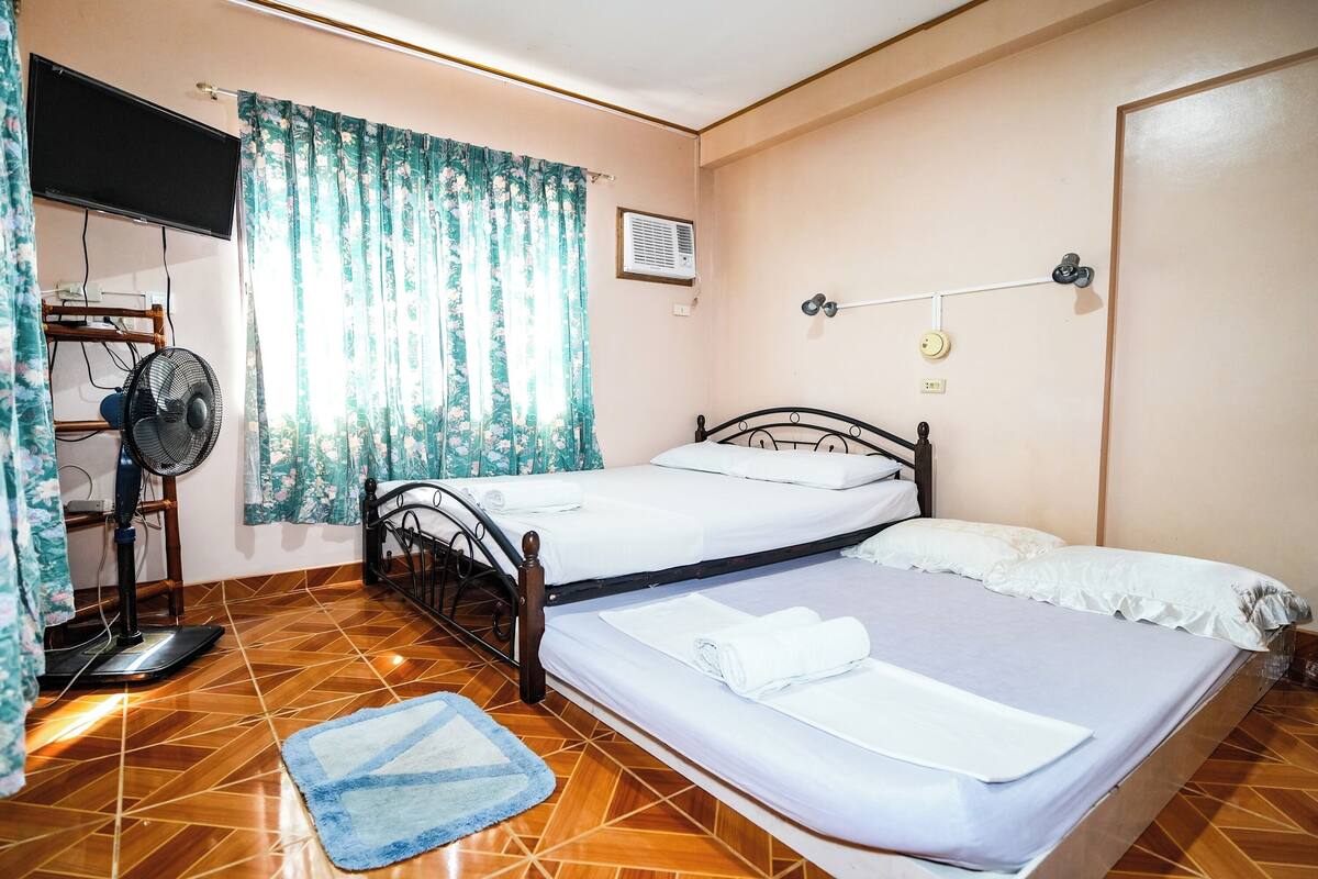 A room with two beds, a standing fan, a TV mounted on the wall, and a window with floral curtains.