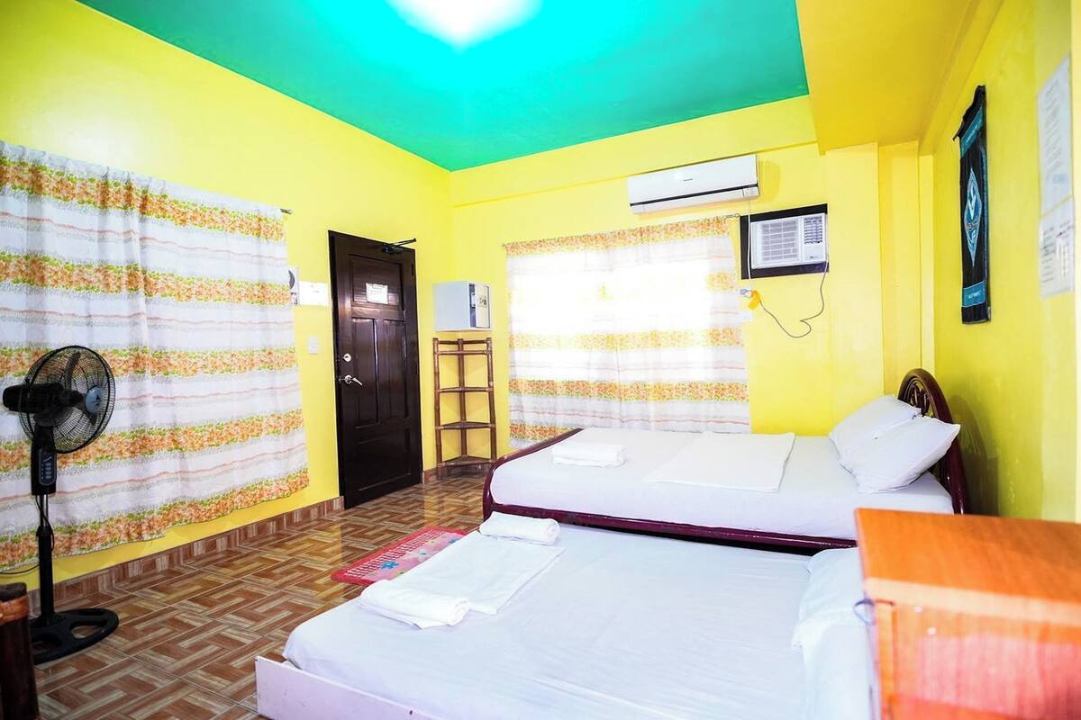 A room with two beds, a standing fan, and an air conditioning unit.