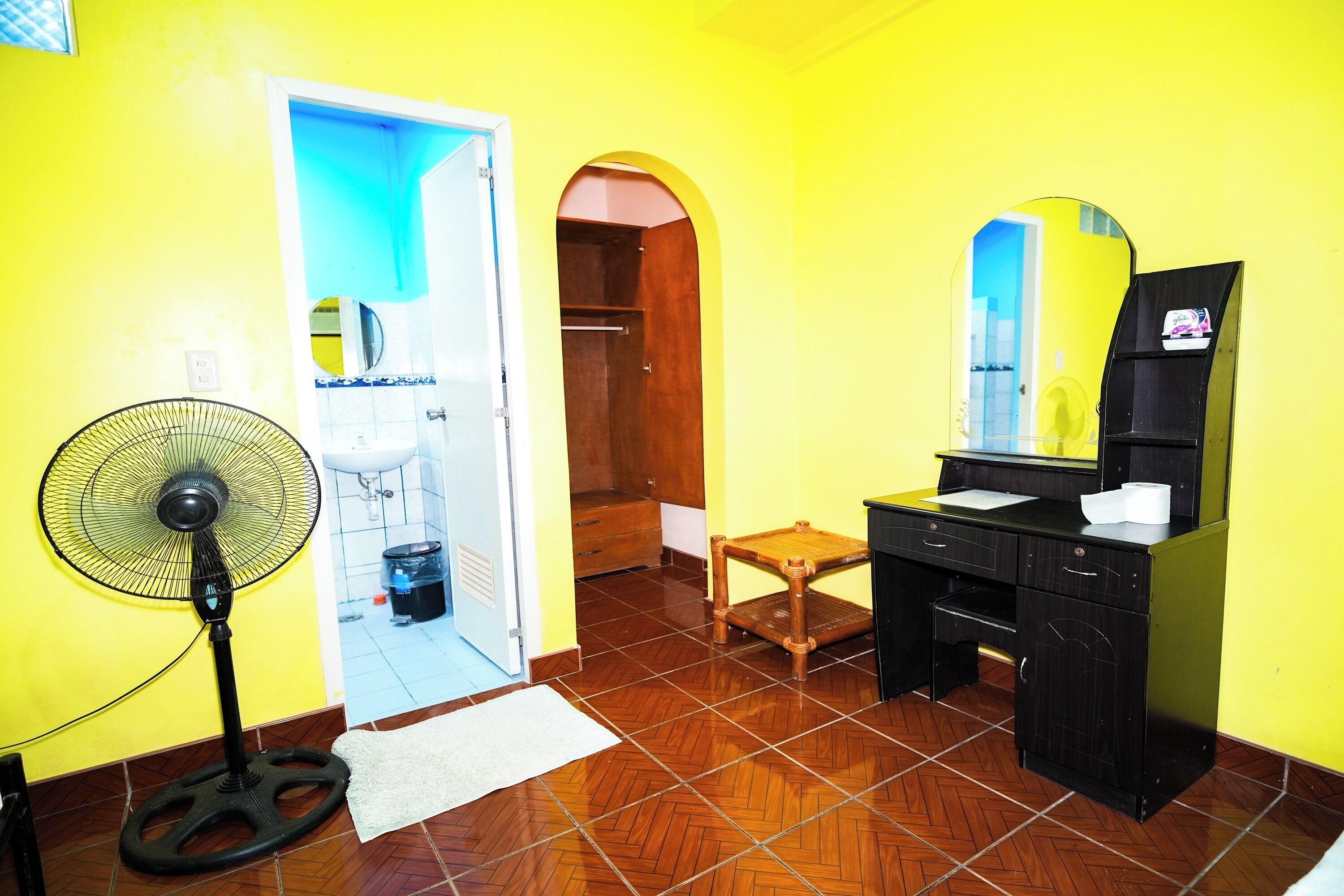 A room with a wooden floor, a fan, a desk with a mirror, and a small stool.