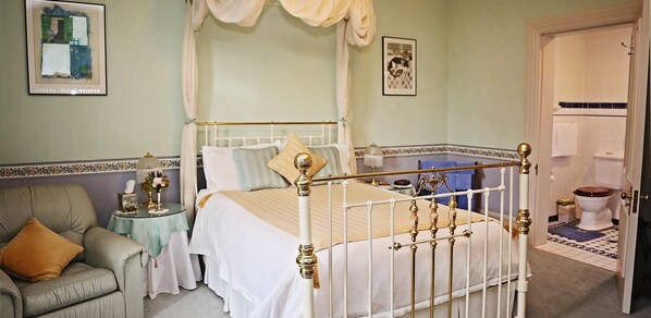 Une chambre à coucher avec un lit à baldaquin, un fauteuil vert, une petite table sur laquelle est posé un vase, et un tableau encadré accroché au mur.
