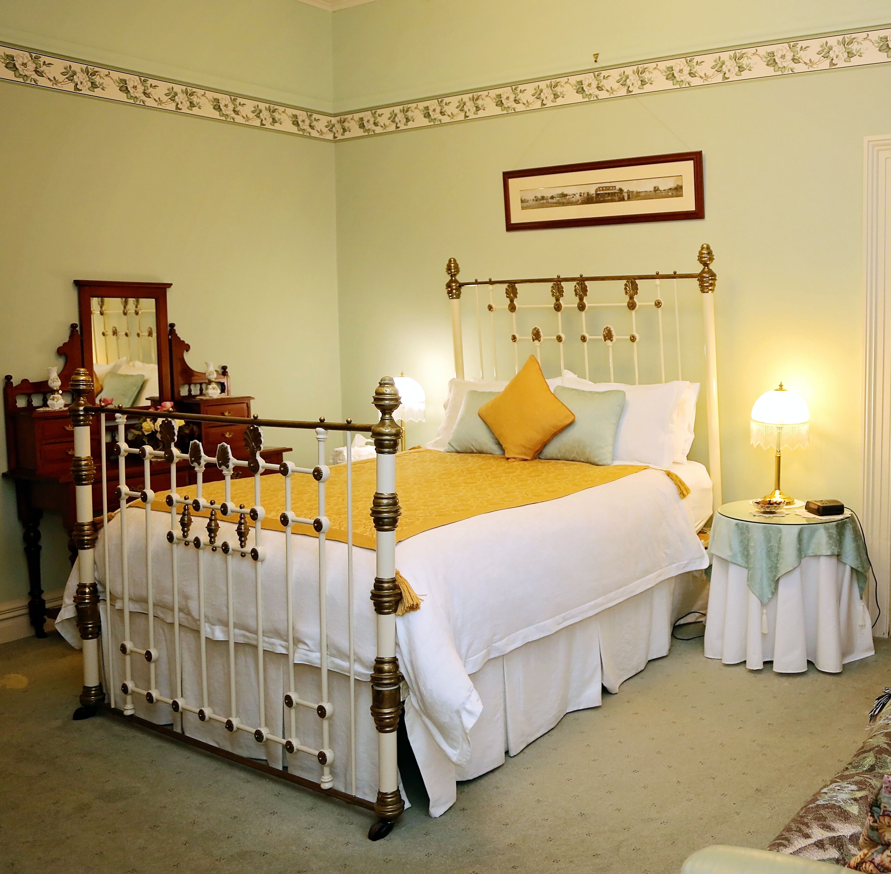 A traditional four-poster bed with a white and gold color scheme, a bedside table with a lamp, and a framed picture on the wall.