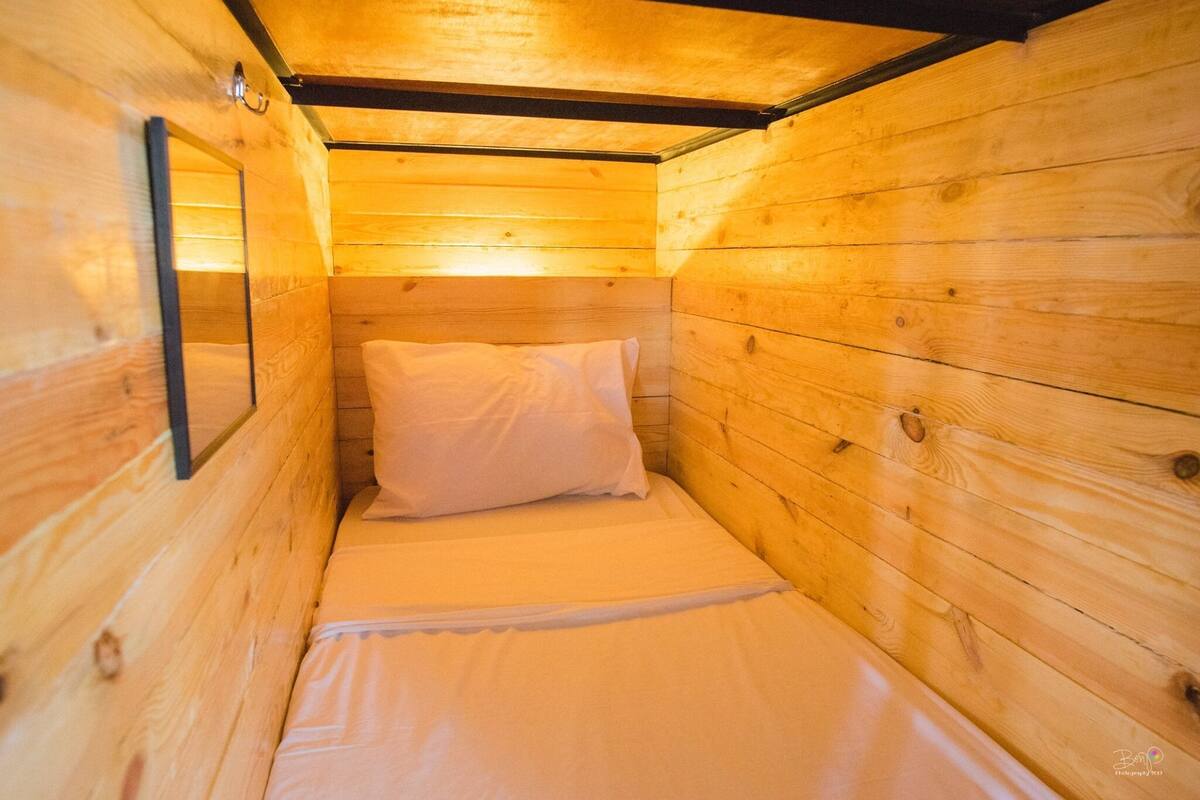 A small, wooden cabin-style room with a single bed, a wooden ceiling, and a wall-mounted light fixture.