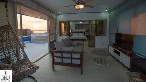 A modern hotel room with a sofa, a TV, a ceiling fan, and a large window offering a view of the sunset. - 3BU Hostel La Union (San Fernando)