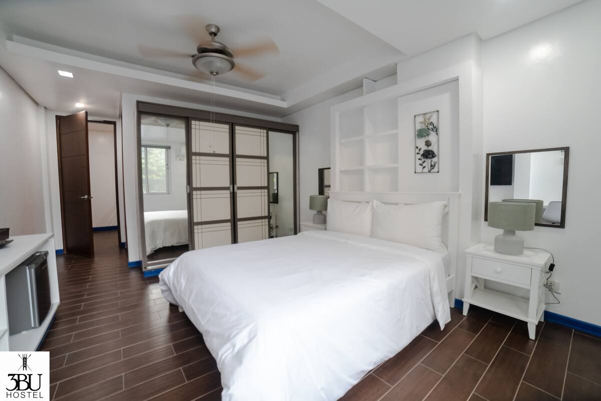 A modern hotel room with a large bed, a ceiling fan, a bedside table with a lamp, and a wardrobe with a sliding door.