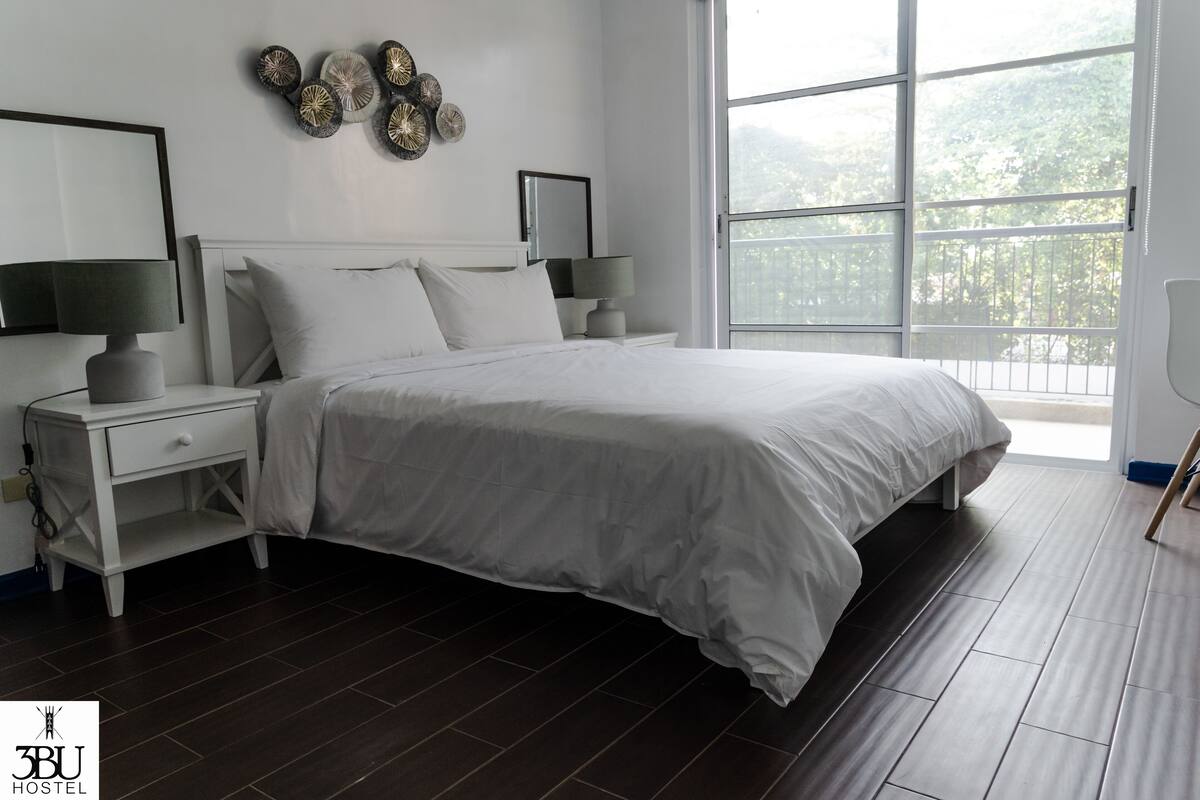 A modern bedroom with a large bed, white nightstands, and a view of greenery through the window.