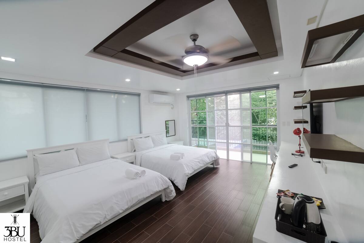 A hotel room with two beds, a ceiling fan, a large window, and a desk with a kettle and a cup.