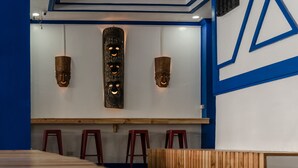 A restaurant interior with a long wooden counter, three red stools, and wall-mounted decorative masks. - 3BU Hostel La Union (San Fernando)