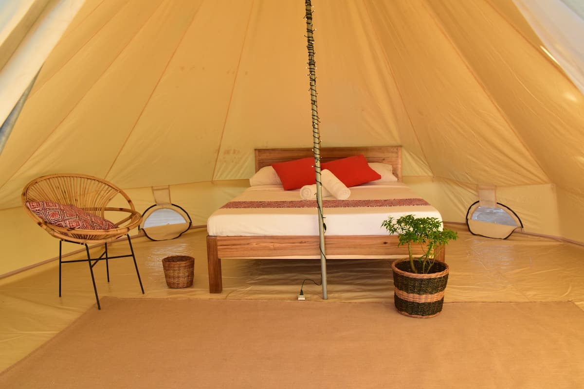 A cozy tent with a bed, two wicker chairs, a small plant, and a hanging light fixture.