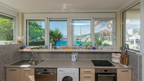 Una cocina moderna con lavavajillas, lavadora y horno, una ventana con vistas a la costa, y una encimera con diversos objetos.