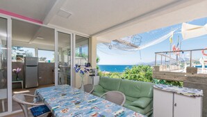 Un balcón con vista al mar, una mesa con mantel colorido, sillas de mimbre, un sofá verde y una pequeña cocina.