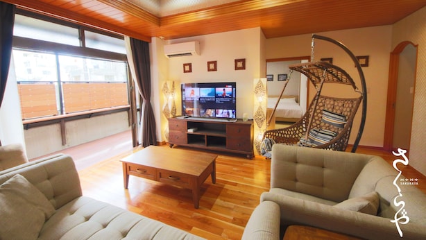 Un salon avec un plafond en bois, un téléviseur posé sur un meuble en bois, un canapé, un fauteuil et une fenêtre avec des stores.