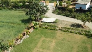 A rural landscape with a pathway leading to a small building with a red roof, surrounded by fields and trees. - Corbett Castle Resort (Kaladhungi)