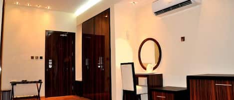 A hotel room with a wooden floor, a desk with a chair, a mirror, and a wardrobe.