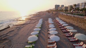 A beach with rows of sun loungers and umbrellas, a stone wall, and buildings in the background.