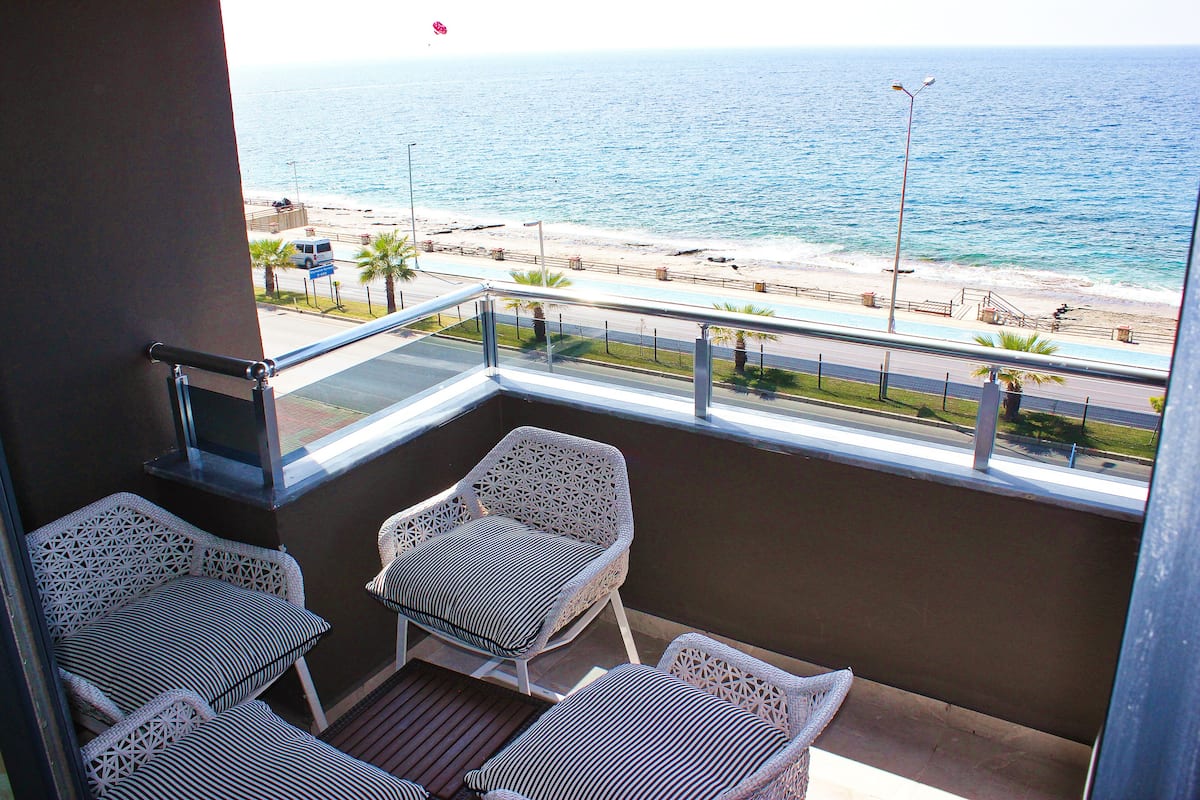 Un balcon avec des fauteuils en osier et une petite table, donnant sur une plage bordée d’une route et de palmiers.