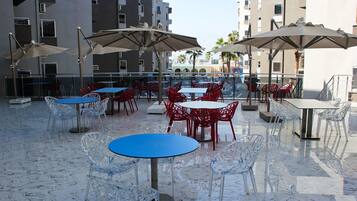 Un patio al aire libre con mesas y sillas, algunas con sombrillas, y una piscina al fondo.