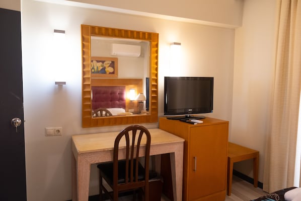 Habitación de hotel con un escritorio de madera, un televisor en un mueble, un espejo y una lámpara.