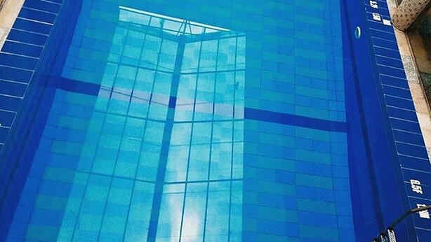 A view of a swimming pool with clear blue tiles and a glass ceiling, reflecting the sky and surrounding buildings. - Carabana Hotel & Suites (Uyo)
