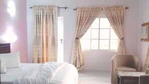 A bedroom with a bed, a chair, a small table, and a window with curtains. - Carabana Hotel & Suites (Uyo)