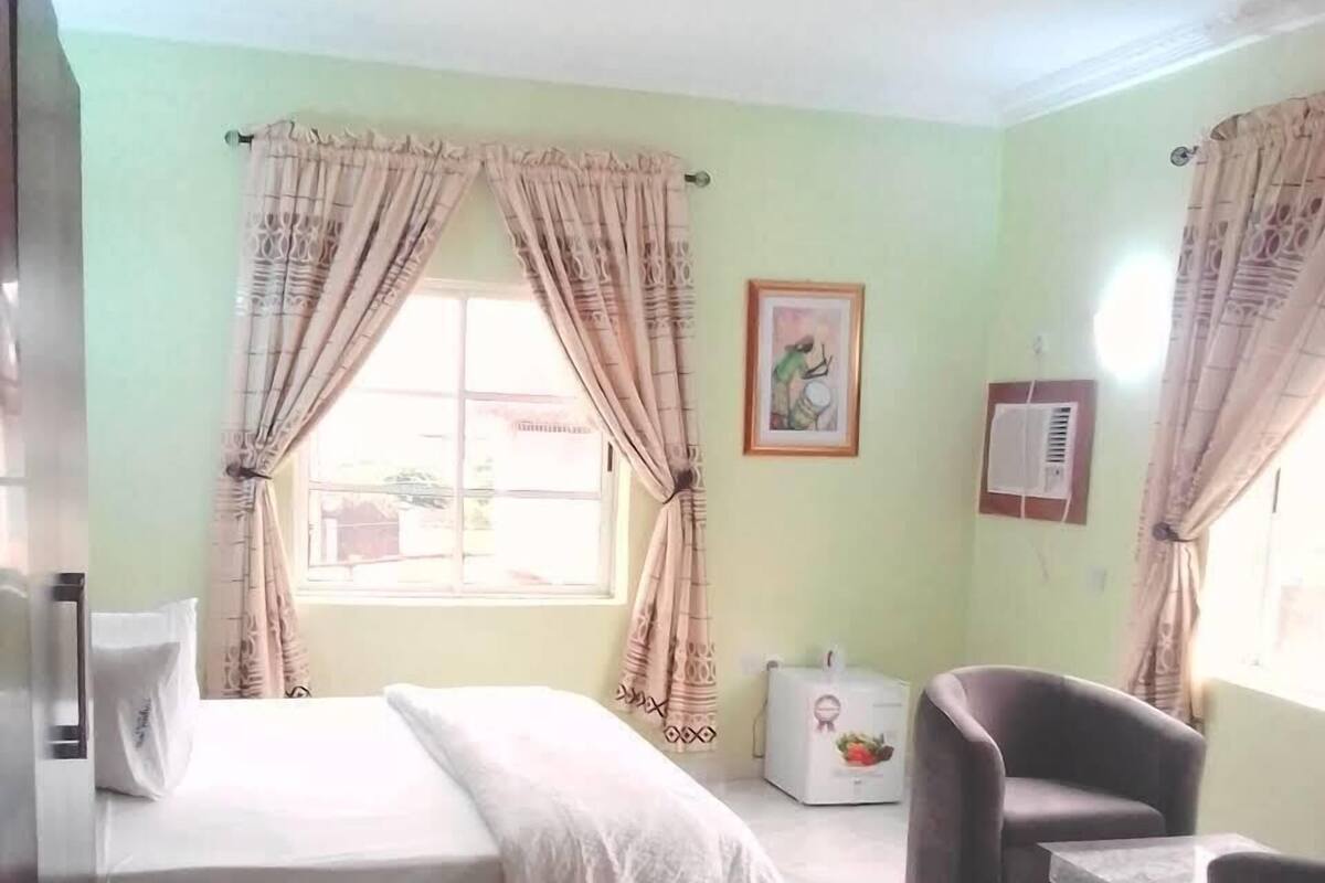 A room with a bed, a chair, a small table, a window with curtains, a picture on the wall, and an air conditioning unit.