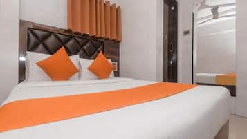 A hotel room with a bed, orange pillows, a white bedspread, a wooden headboard, and a bathroom with a mirror and towel rack.