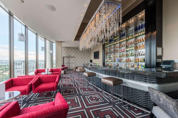 A modern bar area with a high bar and a variety of liquor bottles. There are red seating areas and a patterned carpet. - Southern Sun Sandton (Johannesburg)