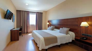 Habitación de hotel con una cama grande, un escritorio, una silla, un televisor montado en la pared y un ventanal con cortinas.