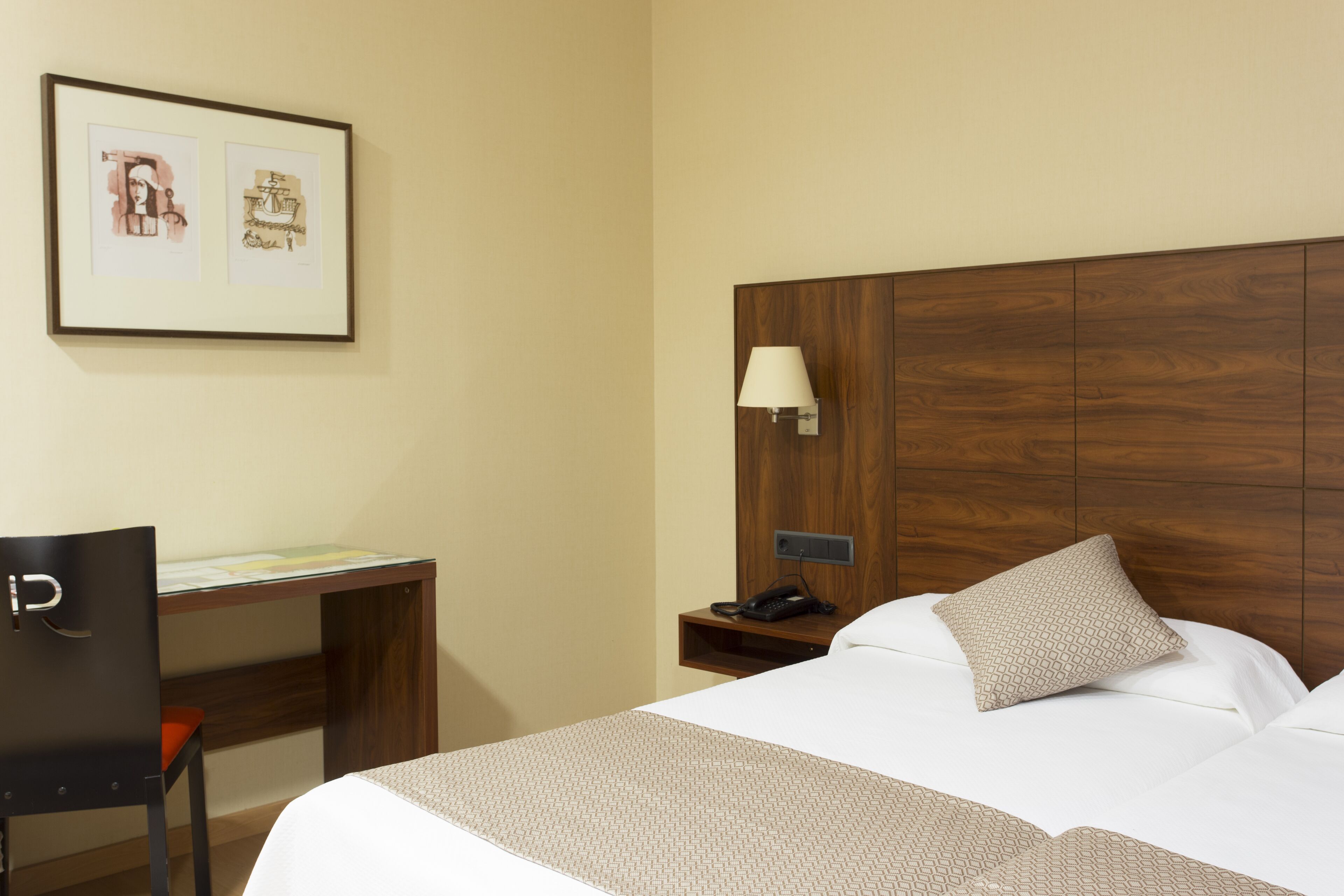 A hotel room with a wooden headboard, a desk with a chair, a bed with a patterned bedspread, a nightstand with a lamp, and two framed pictures on the wall.