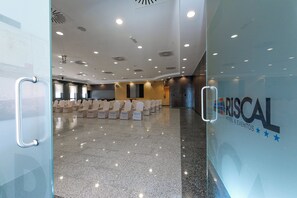 Un salón espacioso con filas de sillas y piso pulido, una pared de vidrio con el logotipo de RISCAL HOTEL & EVENTOS, e iluminación empotrada en el techo.