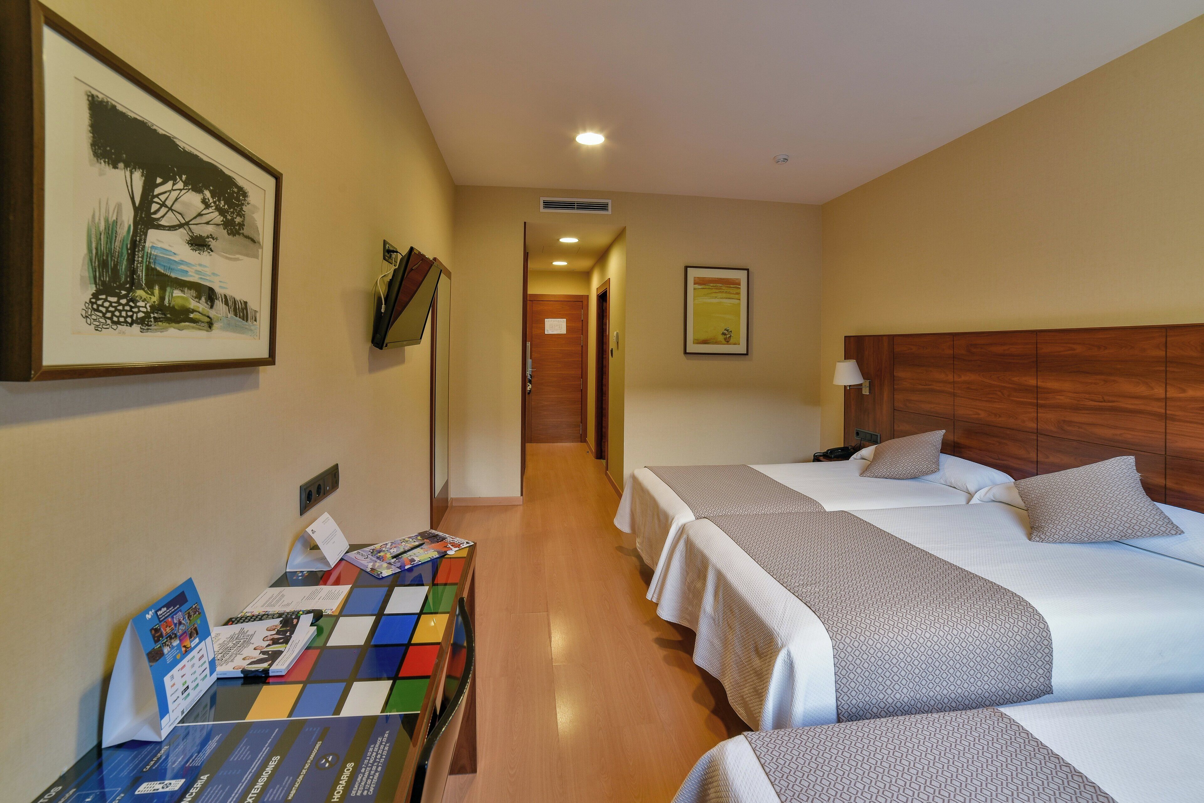 Habitación de hotel con dos camas, un cabecero de madera, un escritorio con un juego de mesa colorido y un cuadro de paisaje en la pared.