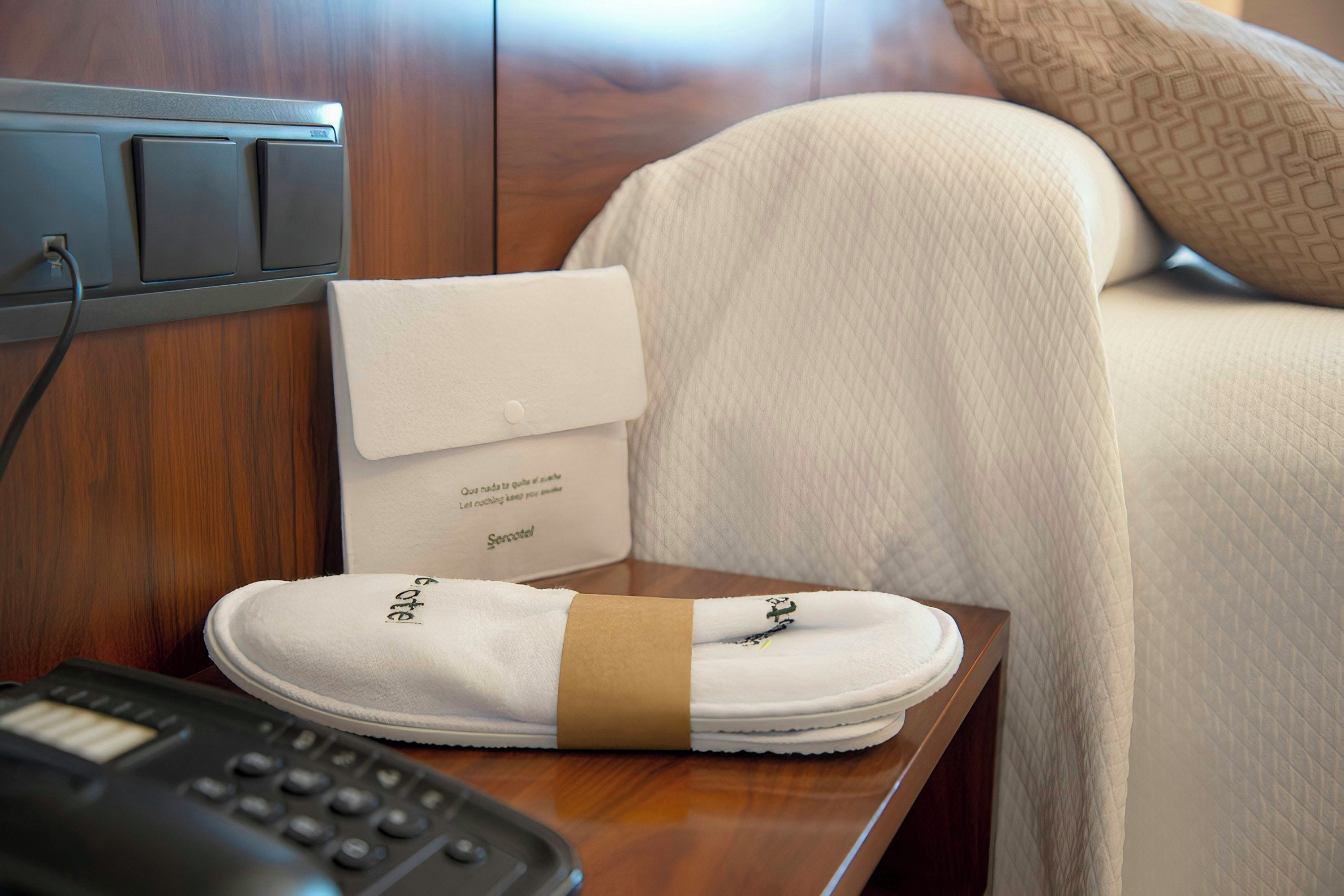 Habitación de hotel con una cama bien hecha, una mesita de noche con teléfono y un cartel que indica los servicios de la habitación.