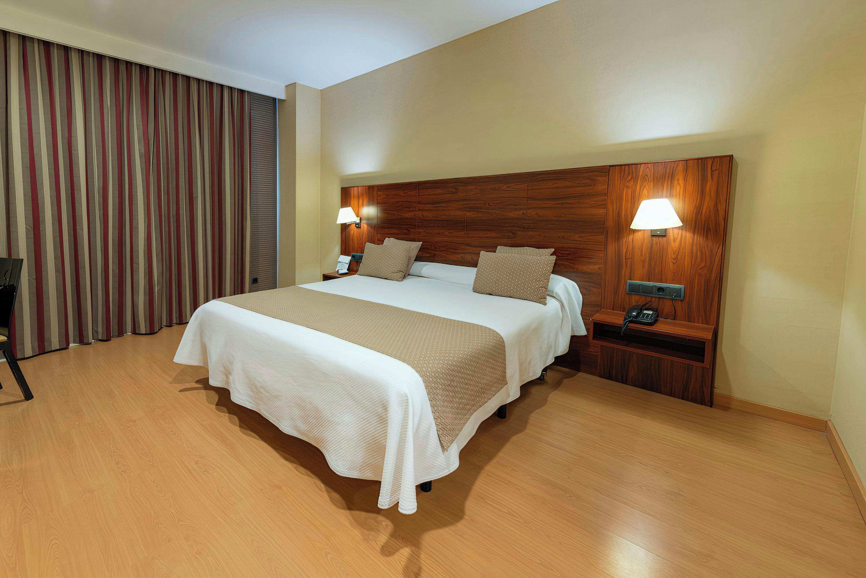 A hotel room with a large bed, wooden headboard, two bedside tables with lamps, a telephone, and a curtain.