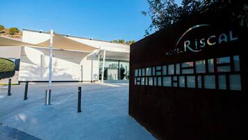 A modern hotel entrance with a clear sky, a distinctive building facade, and a sign reading 'RISCAL HOTEL'.