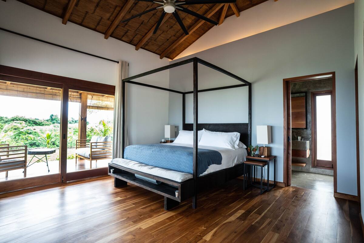 A spacious bedroom with a four-poster bed, wooden flooring, and large windows offering a view of greenery.