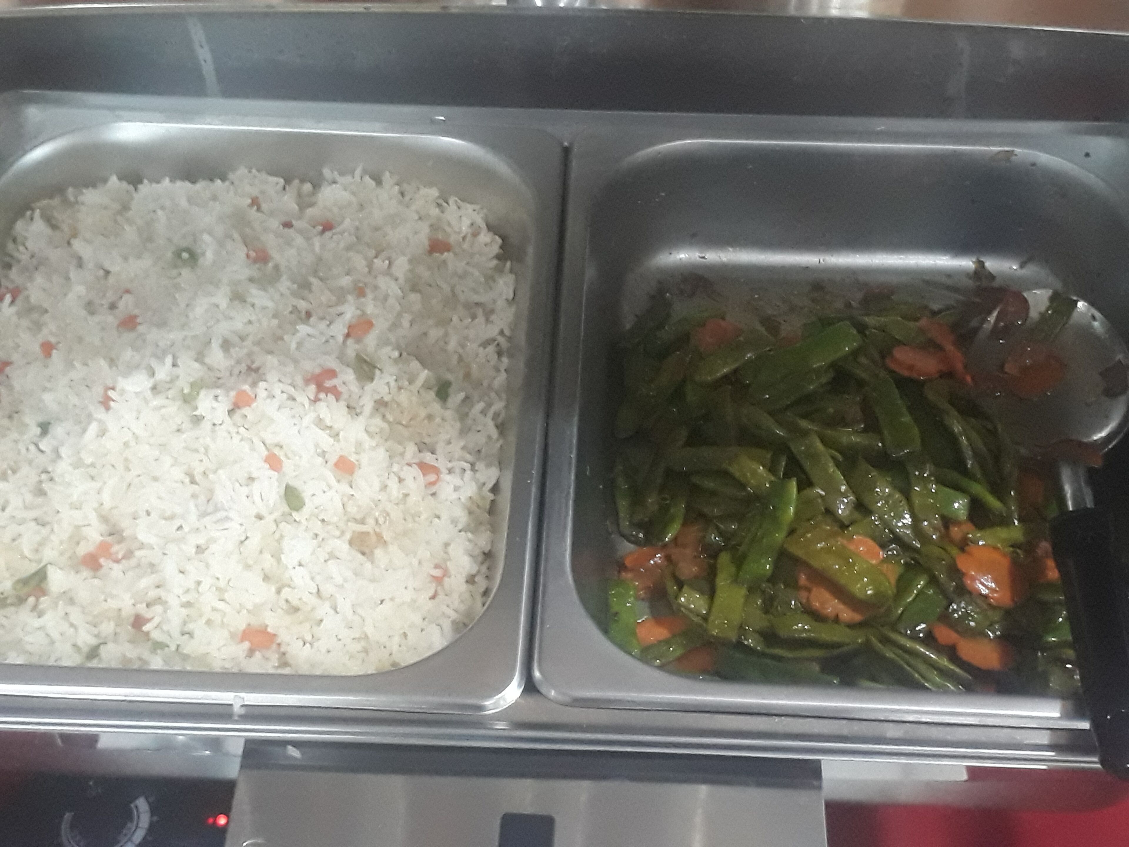 A stainless steel food serving tray with two compartments. One contains white rice with small bits of vegetables, and the other has green beans with carrots.