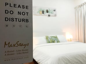 A hotel room with a bed, a nightstand, and a lamp. There is a sign on the wall that reads "PLEASE DO NOT DISTURB" with Chinese characters below it. The room has a white color scheme and a window with curtains.
