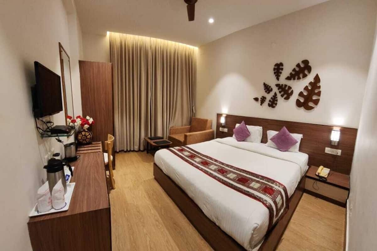 A hotel room with a bed, bedside tables, a desk with a flat-screen TV, a chair, a wardrobe, and a large window with curtains.