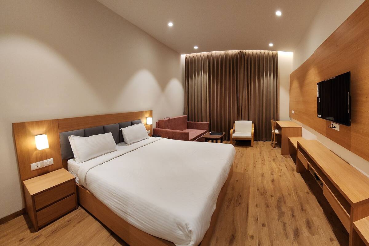 A modern hotel room with a large bed, wooden headboard, bedside tables, a flat-screen TV mounted on a wooden panel, a seating area with a chair and a small table, and a large window with curtains.