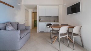 Ein kompakter Wohnraum mit grauem Sofa, einem Holzesstisch mit vier Stühlen, einer Küchenzeile mit weißen Schränken und einem an der Wand befestigten Fernseher.